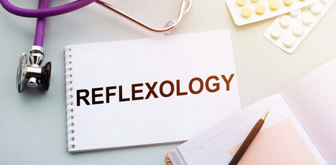 The word REFLEXOLOGY written in notepad with sthethoscope, pills and pen lying on doctor's table.