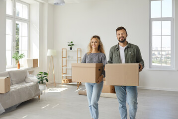 Happy married couple settling down in their new home. Two young people who've bought or moved in a dream house carrying some boxes, unpacking things and making their spacious living room a cozy place