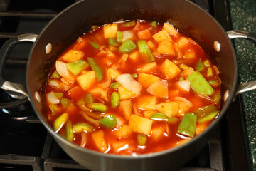 Sweet and sour chicken sauce with green peppers, onion and pineapple chunks in a saucepan on top of...