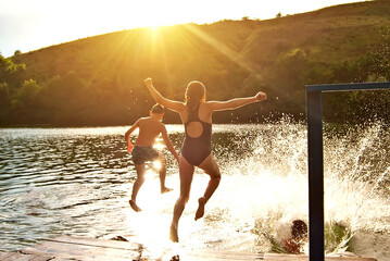 Children jump into the water from the pier. Camping by the water. Active rest on a sunset background. Concept of summer, vacation, travel and vacation. Water splashes on a background of sunlight.