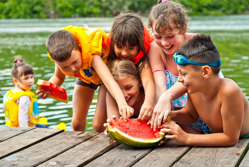 Children eat watermelon on the pier. Children in life jackets rest on the river bank. Camping by the water. - obrazy, fototapety, plakaty