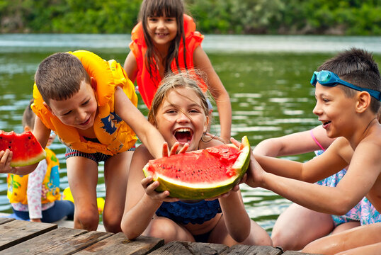 Children Eat Watermelon On The Pier. Children In Life Jackets Rest On The River Bank. Camping By The Water.