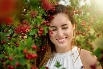 A beautiful young girl with long brown hair and brown eyes against a background of a green tree with bright red berries. Autumn portrait of a girl with her eyes closed and smiling beautifully