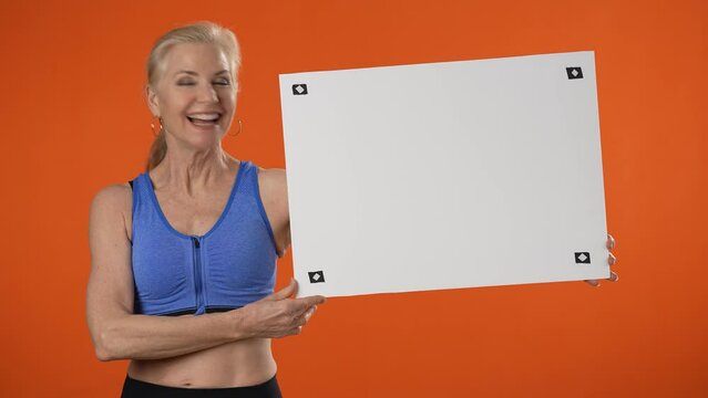 Mature strong sporty athletic fitness woman hold white blank sign board with place for text show muscles isolated on plain orange background