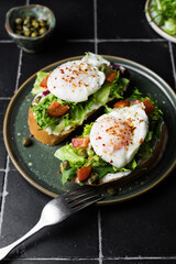 Toast with poached egg, lettuce, tomato and capers on a dark background, close up