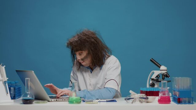 Insane foolish female chemist using laptop and beaker in front of camera, acting crazy and amusing. Mad funny woman with messy wacky hair looking goofy and funny, being silly in studio.