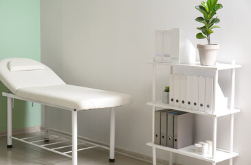 Shelving unit with folders, houseplant and medical couch near light wall