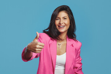 Cheerful young business woman dressed in stylish pink suit showing thumb up while standing and looking at camera against blue background