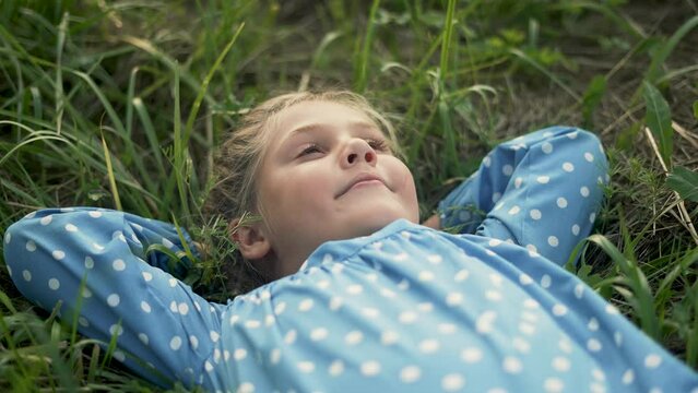 Happy little girl lies on green grass. The child is dream on the grass in park. Happy face of baby. Little girl looks at sky. Smile of baby. Girl is dream lies on grass. Happy baby face look up to sky