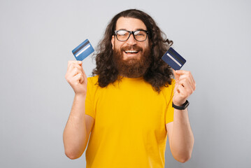 Curly bearded hipster is holding two credit cards over grey bakcground.