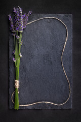 Top view of a black slate board with a bunch of lavender flowers tied with a brown string, which is placed around as a frame. Romantic theme with copyspace for your text.