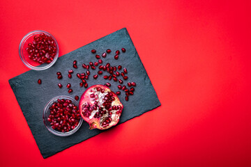 Detail of the fresh pomegranate seeds and juicy pomegranate fruit on the dark slate plate on the red background with empty copyspace.