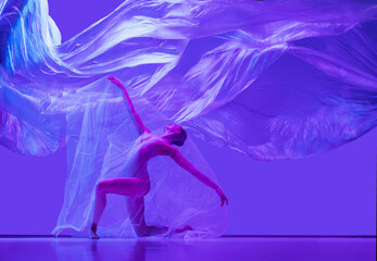 Obraz na płótnie Canvas Portrait of beautiful flexible woman, ballerina dancing with cloth on purple studio background in neon. Fashion, style, art, beauty