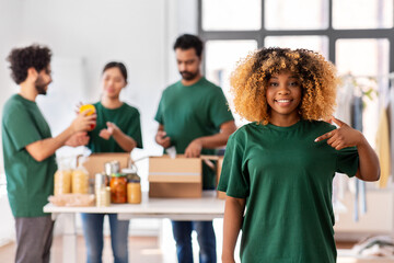 charity, donation and volunteering concept - happy smiling female volunteer pointing to herself and international group of people packing food in boxes at distribution or refugee assistance center