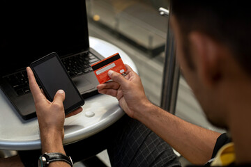 Young male (22) Latin American sitting holding a credit card in his hand and using a cell phone to make Internet payments, digital banking and online shopping, e-commerce concept.