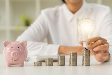 Young woman holding a light bulb next to a piggy bank on a pile of coins on the table, saving money...