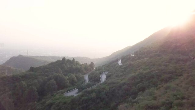 Aerial Flying Over Winding Roads Near Daman E Koh In Margalla Hills With Sun Poking Through Hillside 
