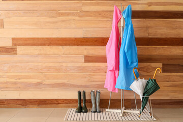 Rack with raincoats, gumboots and umbrellas near wooden wall