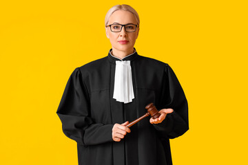 Mature female judge in robe with gavel on yellow background