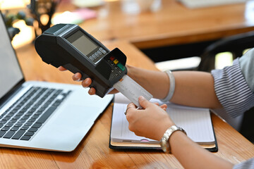 Cropped view of a woman sitting in the office holding a credit card and using a credit card swipe machine, for finance and technology concept.