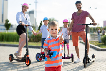 Happy family rides electric scooters and gyroscuters in the park, in the foreground a small boy holds a skate in his hands and gives a thumbs up