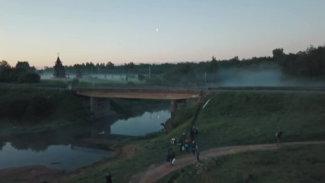 Young people on a lonely road on a bridge near a church and river on a foggy morning at dawn. River, forest, fog, village