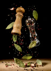 Fototapeten Two spice mills with peppercorns and laurel bay leaves on black flying over table. © Soho A studio