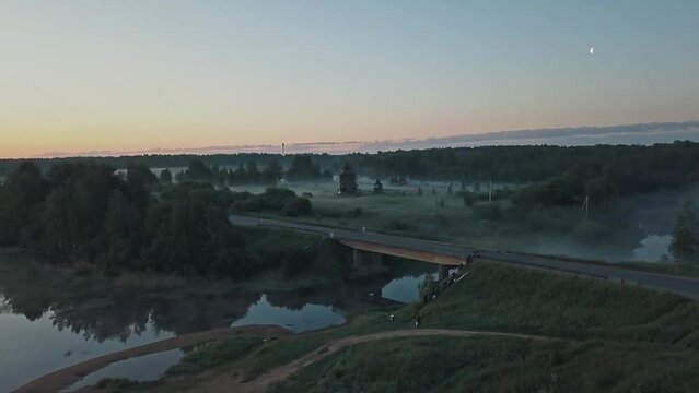Young people on a lonely road on a bridge near a church and river on a foggy morning at dawn. River, forest, fog, village