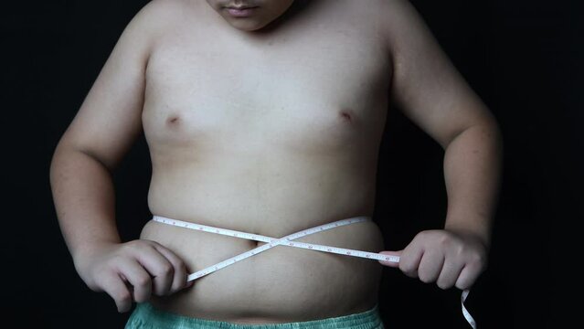 Obese boy measures his fat belly with a measuring tape on black background, health care and diet food concept