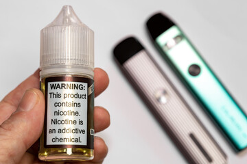 Electronic cigarette liquid selective focus. On the plastic bottle it says "Warning: This product contains nicotine. Nicotine is an addictive chemical." Nicotine vapor sticks are out of focus.