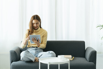 Obraz premium Smiling pretty asian woman in sweater using tablet computer while sitting on sofa at home.