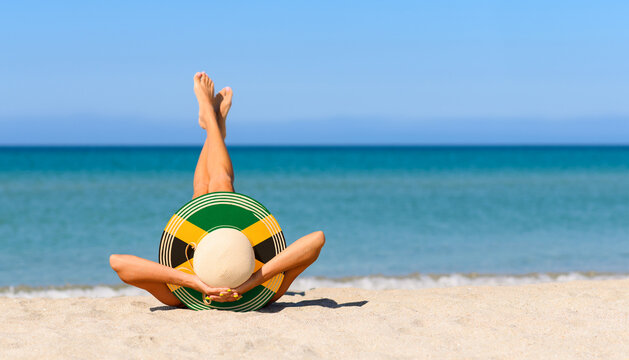 A slender girl on the beach in a straw hat in the colors of the Jamaican flag. The concept of a perfect vacation in a resort in the Jamaica. Focus on the hat.