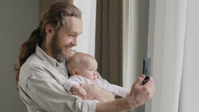 Happy smiling caucasian father adult daddy holding newborn child at home standing near window dad with little daughter son infant newborn making photo video vlog online stream conference distant call