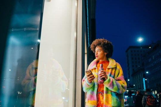 Young mixed race woman using reusable water bottle and smartphone walking outdoor at night