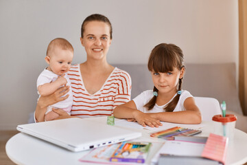 Indoor shot of positive optimistic young mother sitting with schoolgirl and infant daughters at table, doing school tasks at home, looking at camera.