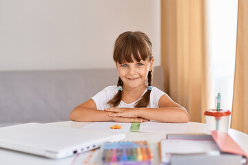 Naklejka na ściany i meble Portrait of happy satisfied child girl doing homework writing and reading at home, cute female child with dark hair and braids looking at camera with positive expression, distance learning.