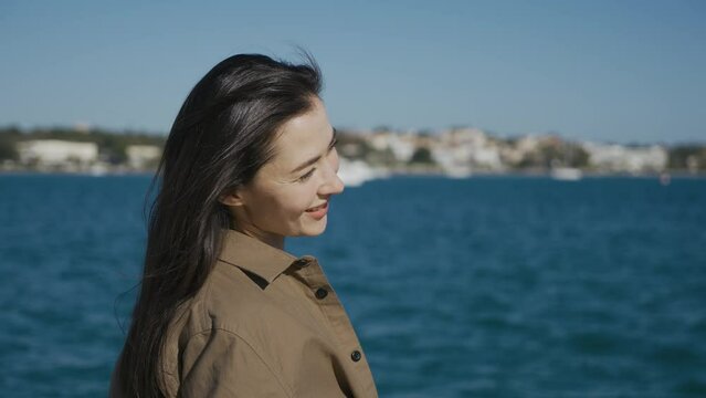 A girl, a brunette, of Asian appearance, is standing on a pier on the seashore, smiling. Closes eyes. The wind is blowing, the sun is shining. Close-up.