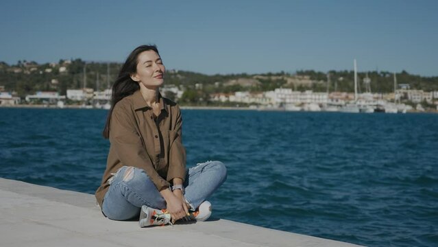 A girl, a brunette, of Asian appearance, sits on a pier on the seashore, touches her hair, smiles. The wind is blowing, the sun is shining.