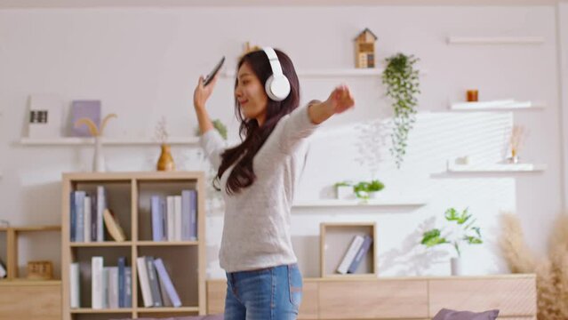 Happy Young women listening to music in headphones and dancing energetic and rhythm at home. Attractive Cheeful Asian girl looking at the smart phone sitting in the living room spending leisure time.