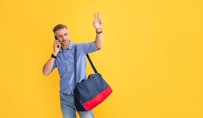 man speaking on smartphone with travel bag gesturing hello on yellow background, copy space, peace