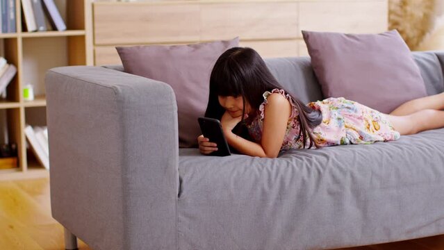 Cute little girl uses smartphone while prone at the sofa in the living room. Child surfing the internet on computer, browses through internet and watches cartoons online.