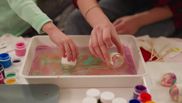 Mother and girl roll eggs in tray with paints and oily water