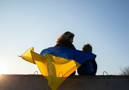 little boy and a woman sit with their backs at sunset against the sky with a blue and yellow Ukrainian flag. Family, refugees, unity, support. Ukrainians are against the war. stand with Ukraine