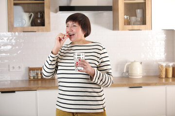 Happy middle aged 50s woman holding pill and glass of water taking dietary supplements. Portrait of smiling adult attractive woman taking care of health in menopause, at home.