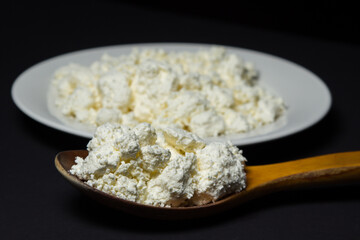 A plate of cottage cheese on a black background. Healthy and tasty dairy product. Wooden spoon full of cottage cheese
