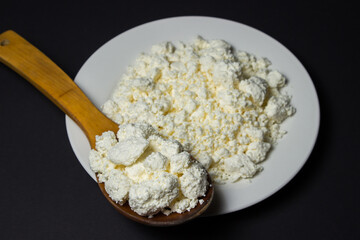 A plate of cottage cheese on a black background. Healthy and tasty dairy product. Wooden spoon full of cottage cheese