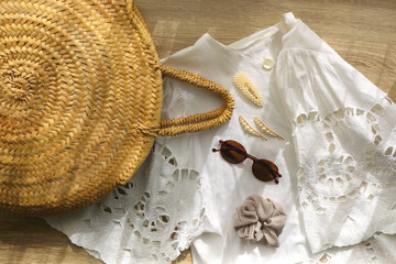 White linen lace shirt, round straw bag, sunglasses and cute hair accessories on wooden background....