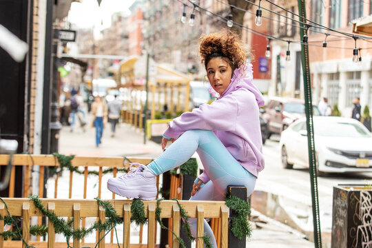 Young woman with black and brown curly hair wearing a pink hoodie and blue yoga pants sits outside in city