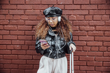 Young woman wearing black leather jacket and beret listening to music on headphones with suitcase for travel next to brick wall - obrazy, fototapety, plakaty