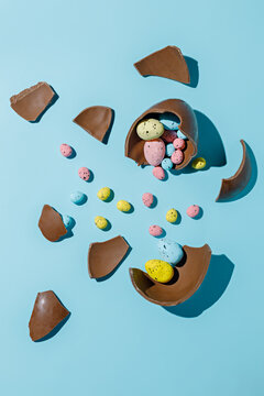 Easter chocolate egg broken in pieces with small multicolor eggs inside on pastel blue background. Creative Easter holiday or sweeties concept. Minimal festive greeting card.